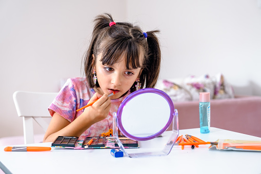 little girl applying make-up and looking at her reflection in mirror while sitting at the dressing table