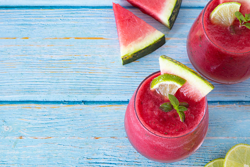 Watermelon smoothie drink in a glass decorated with fresh wedges, lime, and mint leaves on a blue wooden table with copy space. Top view. Cold and refreshing summer juice.