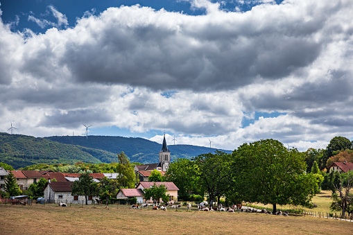 Village with church steeple, agriculture with some cows on foreground and wind turbines in background on mountain ridge. This image was taken in Chamagnat, Saint-Alban near Cerdon small village of Bugey, Alps mountains near Jura massif, in Ain, Auvergne-Rhone-Alpes region in France.