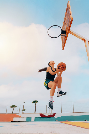 Low angle of woman playing basketball and jumping hoop against blue sky, copy space. Street game. Sport, practice and effort