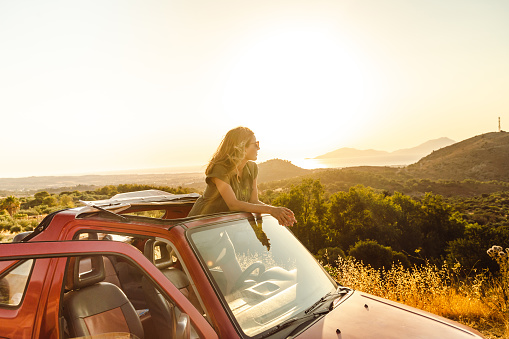 Young woman in an open-top car. She is enjoying the sunset and looking at the view