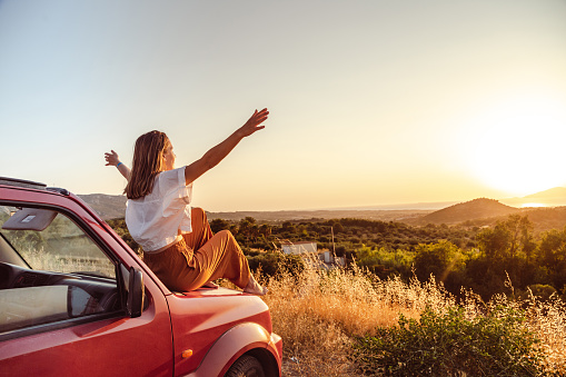 istock Young woman arms raised sitting on the car and enjoying the sunset 1412545366