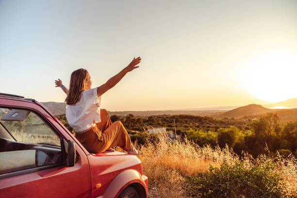 młoda kobieta z podniesionymi rękami siedzi na samochodzie i cieszy się zachodem słońca - road landscape journey road trip zdjęcia i obrazy z banku zdjęć