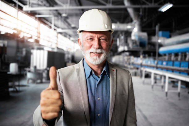 Portrait of successful senior factory manager in business suit and hardhat holding thumbs up in industrial production hall. stock photo