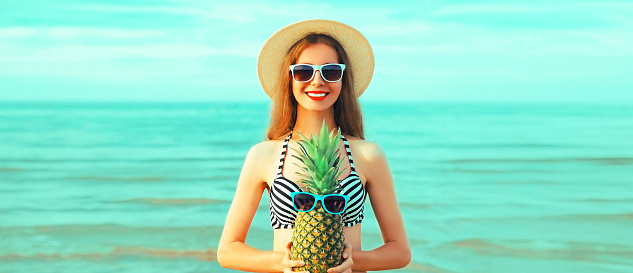Portrait of happy smiling young woman with pineapple wearing straw hat on the beach on sea background at summer day