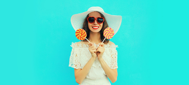 Portrait of happy smiling young woman with sweet lollipop wearing red heart shaped sunglasses wearing summer white straw hat on blue background