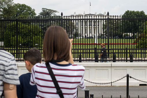 Washington, DC, USA - June 22, 2022: A worker walks past the White House, the presidential residence and workplace in Washington, DC, enclosed by tall fences. Tourists are seen in the blurry foreground.