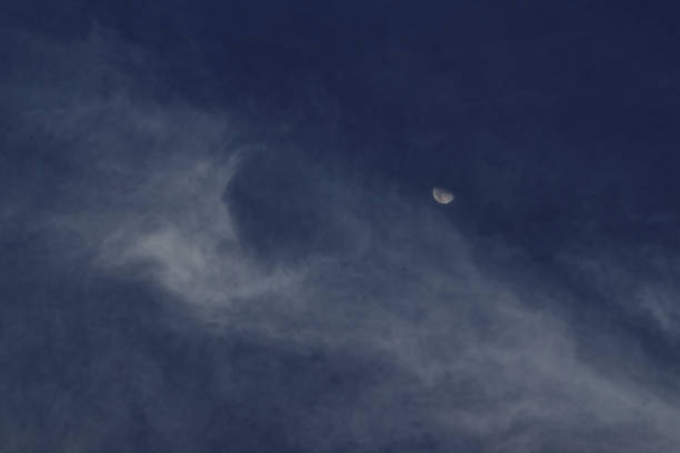 thin veil of white clouds blown by the wind, veiling a distant crescent moon under a blue sky - POA, SAO PAULO, BRAZIL.