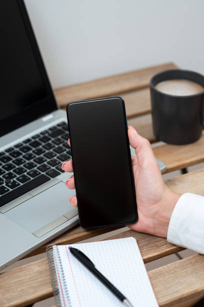 istock Businesswoman Holding Cellphone With Important Message On Table With Lap Top,Coffee, Notebook And Pen. Sitting Woman Presenting Crutial Information On Paper. 1412562559