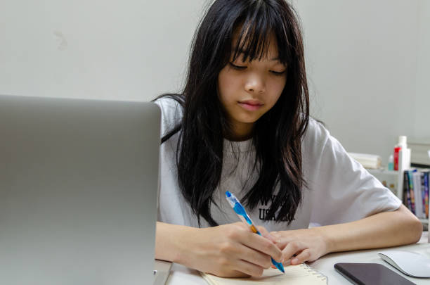 istock girl holding pen writing book study learning digital internet video online computer laptop at home. 1412563676