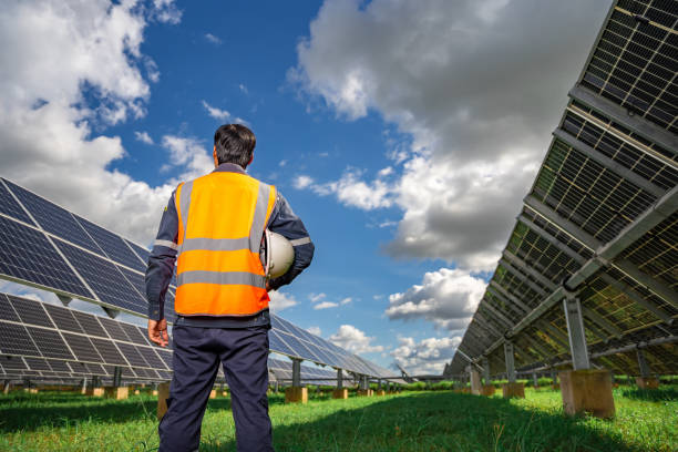 istock Technician dressed in blue uniform and in an orange reflective vest and holding a safety helmet stands facing a solar panel at solar power plant, Photovoltaic module idea for clean energy production 1412564097