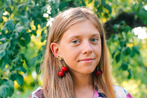 Defocus beautiful blond teen girl with sweet cherry on ear. Beautiful smiling teenage girl closeup, against green of summer park. Child earrings.  Out of focus.