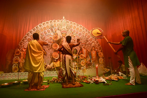 Howrah, India -October 13, 2021 : Hindu Priests worshipping Goddess Durga with mirror, ghanta, chamor and hand fan. Ashtami puja aarati - sacred Durga Puja ritual - shot at night.