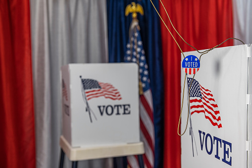 An opened metal coat hanger with an “I VOTED TODAY” sticker hanging on a voting booth with red, white and blue bunting and an American flag in the background of this voting precinct.