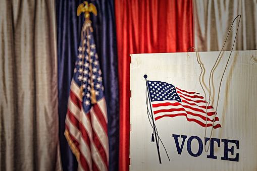 A grungy image of an opened metal coat hanger hanging on a voting booth with red, white and blue bunting and an American flag in the background of this voting precinct.