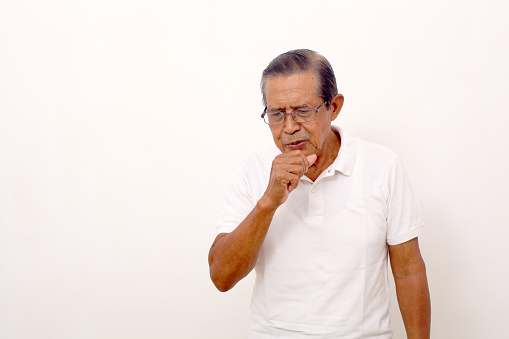 Elderly Asian man standing while coughing over his fist hand. Isolated on white background