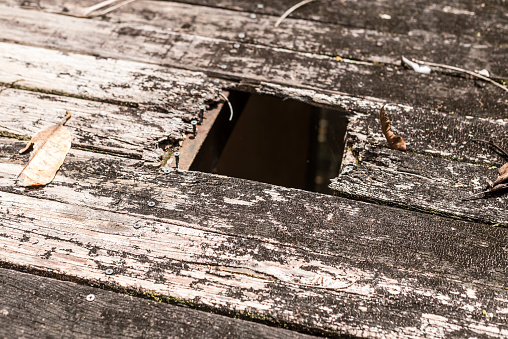 A hole in the wooden board of a 2nd floor deck, posing a hazard. Old and rotten wood planks due to exposure to weather.