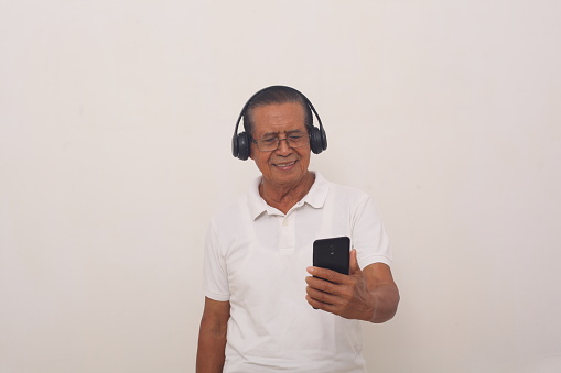 Happy Asian old man standing while listening music from his phone using headphone. Isolated on white