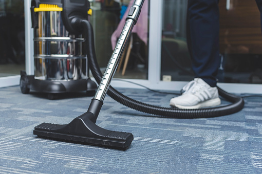 A man cleans the carpet flooring of an office with an upright vacuum cleaner with an attached upholstery nozzle.