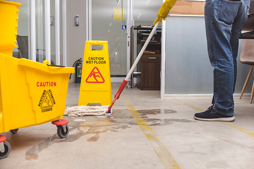 A Caution Wet Floor sign placed to warn passersby while wiping a wet concrete floor with a string mop.