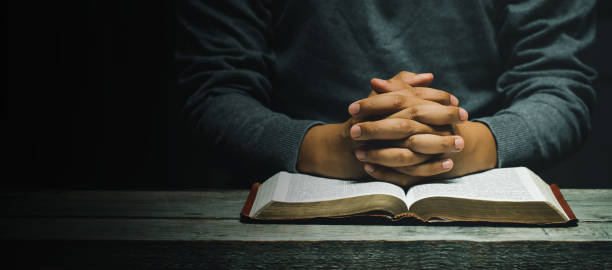 hand of man while praying for the christian religion, man praying with his hands together with bible on a wooden table. begging for forgiveness and believing in goodness. copy space for banner - praying men god kneeling imagens e fotografias de stock