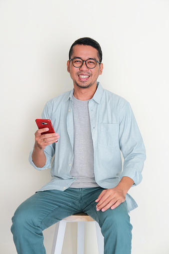 Adult Asian man sitting in a chair holding handphone with happy expression