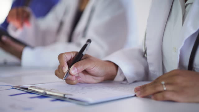 Closeup of a doctor writing notes at a science convention with colleagues and other researchers in the background. Female medical student filling in a clinical trial form to test new medicine