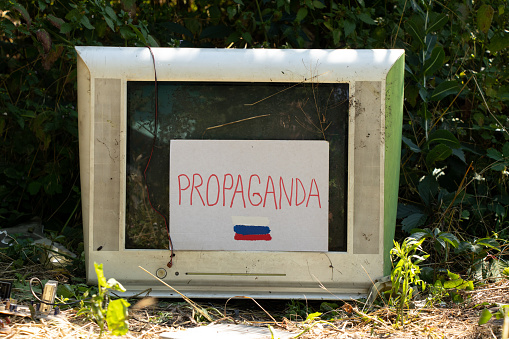 An old broken TV and cardboard paper with text propaganda and a painted Russian flag and the TV itself stands in the forest on the grass in Ukraine..