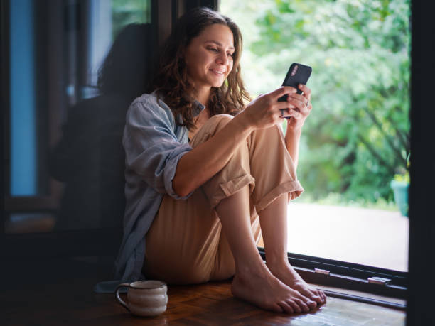 Young beautiful woman with a smartphone sitting on the wooden floor near open door to the terrace of a country house stock photo