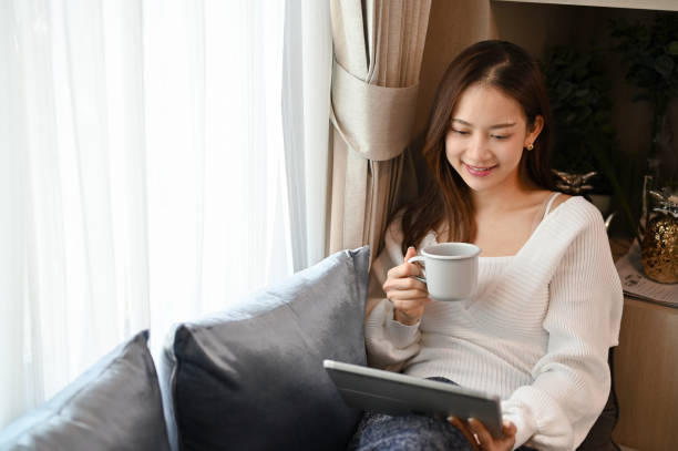 istock Beautiful Asian female chilling in living room, on the sofa, drinking hot coffee while using tablet. 1412617794
