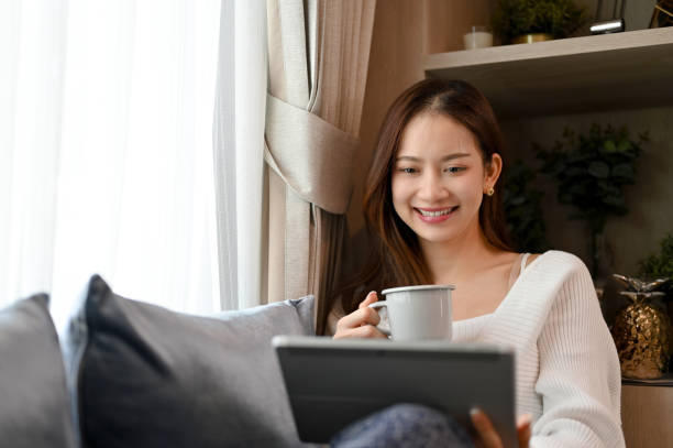istock Happy Asian female chilling in her living room, drinking hot coffee while using digital tablet. 1412617797