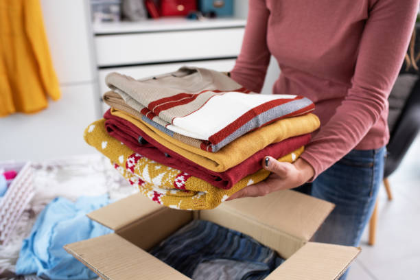 Woman putting her clothes in a delivery box Young woman folding her clothes and packing them in a delivery box, she is changing her wardrobe clothing donation stock pictures, royalty-free photos & images