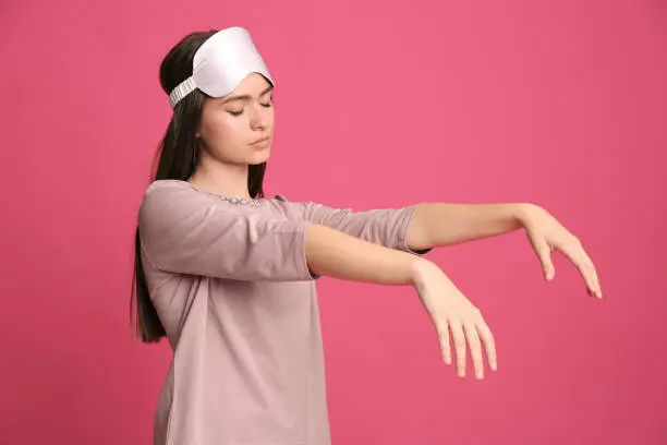 Young woman wearing pajamas and mask in sleepwalking state on pink background