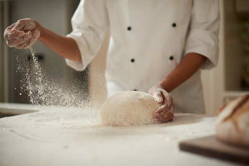 Master chef sprinkling flour over fresh dough on a kitchen table. Closeup hands of bakery chef making and kneading dough for bread using traditional recipe. Enjoying hobby, baking homemade bread