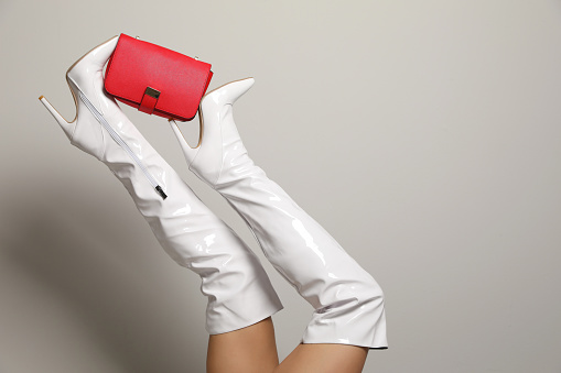 Woman wearing white long shoes with red leather bag on light background, closeup