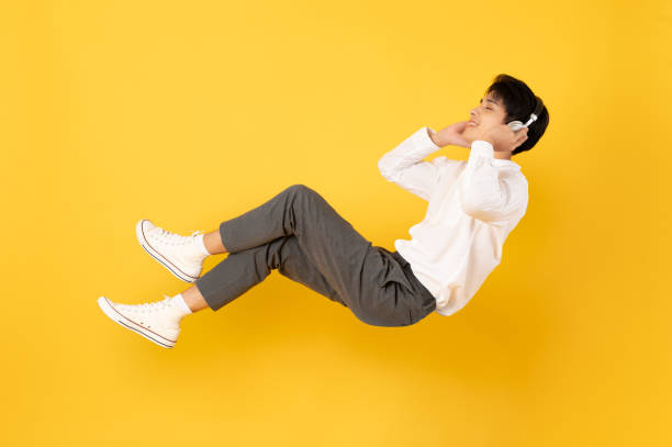 junger, gut aussehender asiatischer mann, der schwebt, während er mit einem isolierten kopfhörer auf gelbem hintergrund zuhört - levitation stock-fotos und bilder