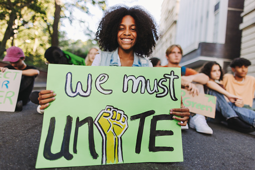 Cheerful black girl holding a poster while sitting with a group of demonstrators at a climate change protest. Multicultural youth activists joining the global climate strike.