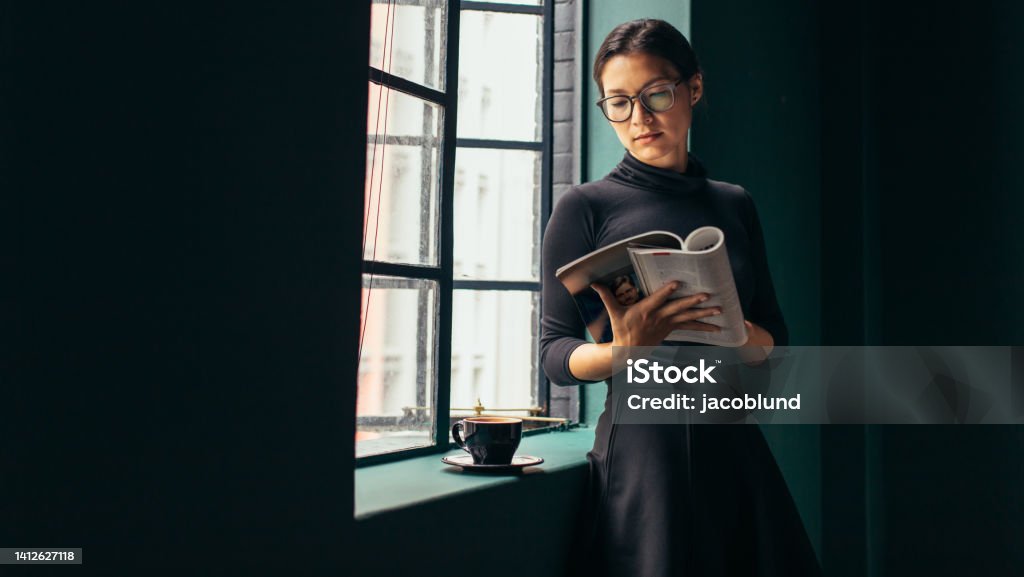 Beautiful female reading fashion magazine Asian woman standing by the window and reading book. Beautiful female reading fashion magazine. Magazine - Publication Stock Photo