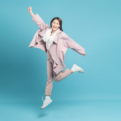 Young beautiful asian woman with smart casual cloth wearing pink coat jumping with excite feeling isolated on blue background