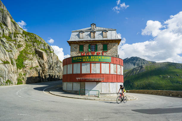 A lone cyclist on the Furka Pass route, riding next to the historic Belvedere Hotel in the Swiss Alps. Furka Pass, Switzerland. 14. August 2021. Historic, closed mountain hotel - Belweder Palace on the furka pass route in the Swiss Alps. furka pass photos stock pictures, royalty-free photos & images