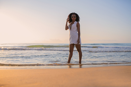 Portrait of an african woman with sunglasses and summer dress standing looking at camera at sunset. Selective focus.