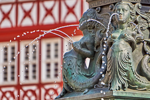 Frankfurt am Main, Germany - July 30, 2022: Water mermaids with splashing breasts at the Justitia fountain on the Römerberg in Frankfurt. Half-timbered in the background. The fountain was built in 1887 as a copy of the 1543 fountain.