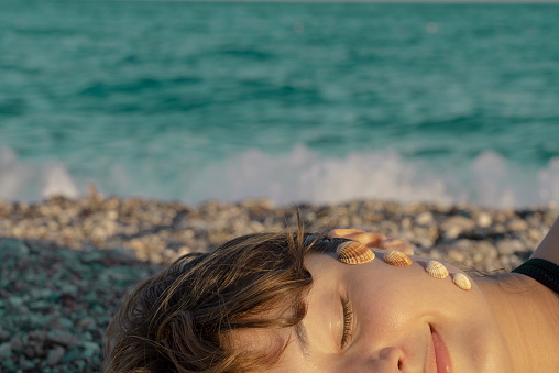Woman posing with seashells on her face.\n\nA woman sunbathing on the beach with seashells on her face.