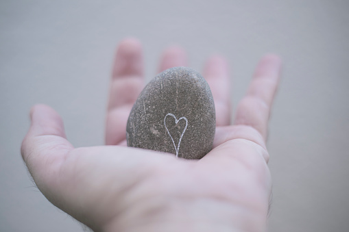 Man's hand holding a gray rounded stone on which there is a white heart drawn in a well-lit environment