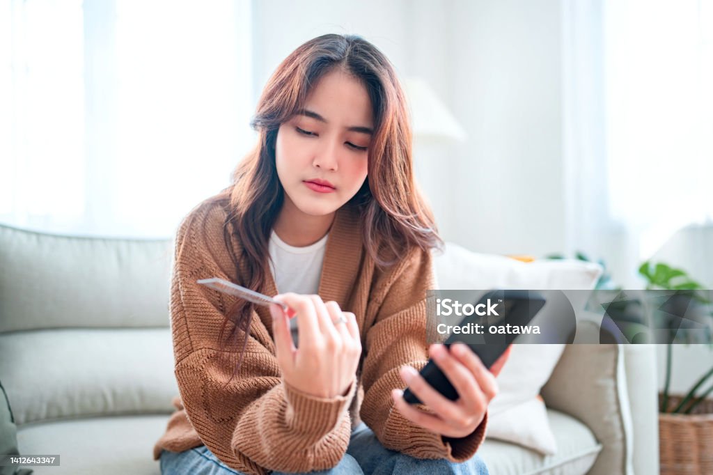Young asian woman problems with paying. Sad female internet shopper sit on couch hold phone and credit card with feeling depressed and worry for distress suffer of overspending money from card account Credit Card Stock Photo