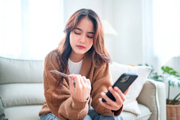 Young asian woman problems with paying. Sad female internet shopper sit on couch hold phone and credit card with feeling depressed and worry for distress suffer of overspending money from card account stock photo