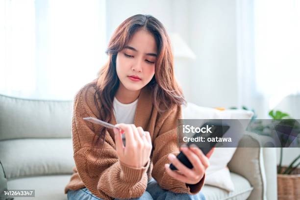 Young Asian Woman Problems With Paying Sad Female Internet Shopper Sit On Couch Hold Phone And Credit Card With Feeling Depressed And Worry For Distress Suffer Of Overspending Money From Card Account Stock Photo - Download Image Now