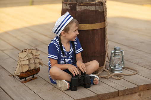A boy in a sailor's uniform against the background of a wooden model of a pirate ship sits near a wooden barrel, next to it is a model of a sailing ship and a kerosene lamp.