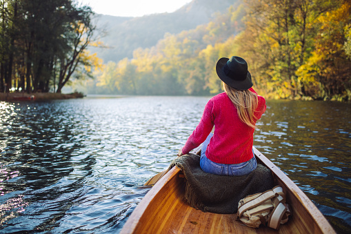Young happy woman enjoying canoeing and paddling on river with nice weather and forest around with autumn colors. She looks beautiful with blond hair. Sunset in background. Photo taken from her back