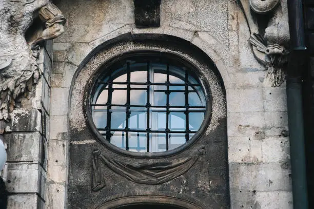 A large round window in a modern metal-plastic frame with a metal lattice on the facade of an old house with stucco and decorative relief elements.